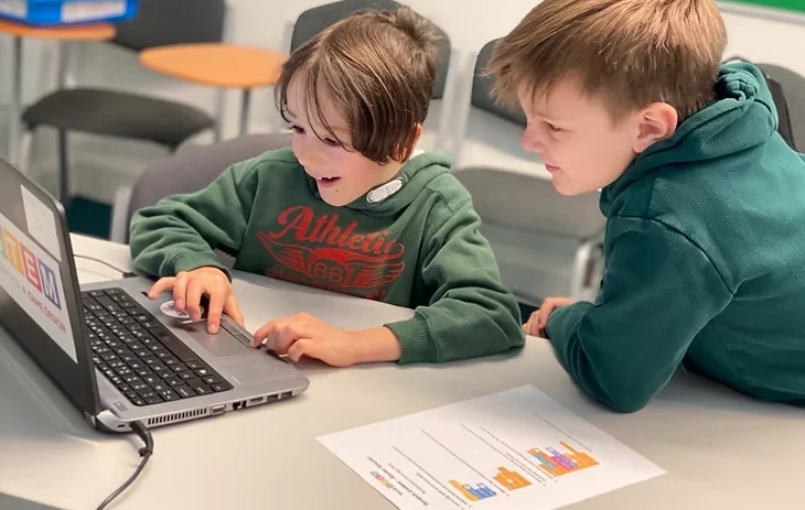 Two boys at a laptop during an AweSTEM club session, one grinning as he works the trackpad while the other watches the screen