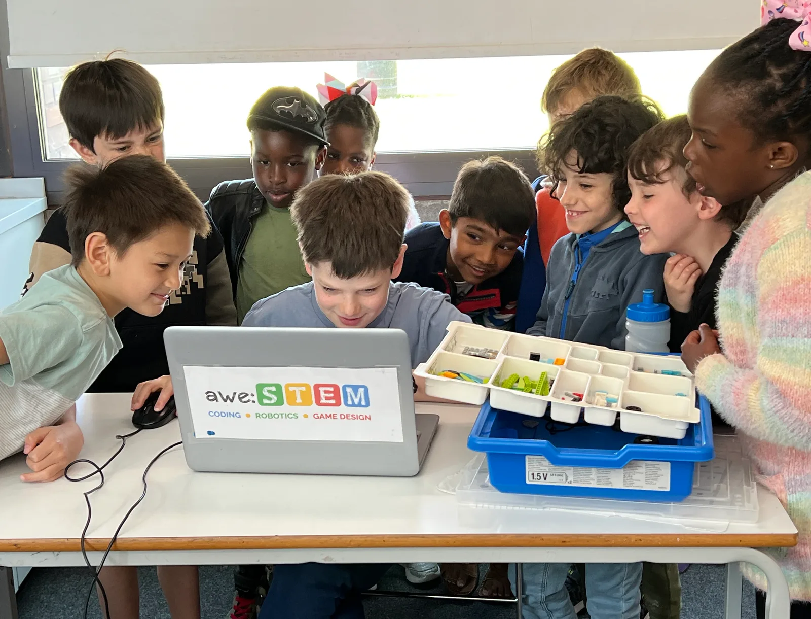 Ten smiling children crowded around an AweSTEM laptop and a tray of LEGO components