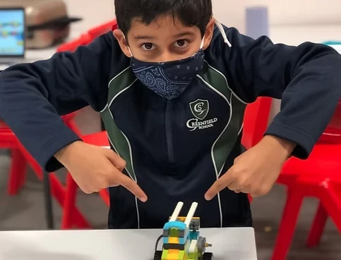 A boy in school uniform pointing proudly at the LEGO robot he has just built