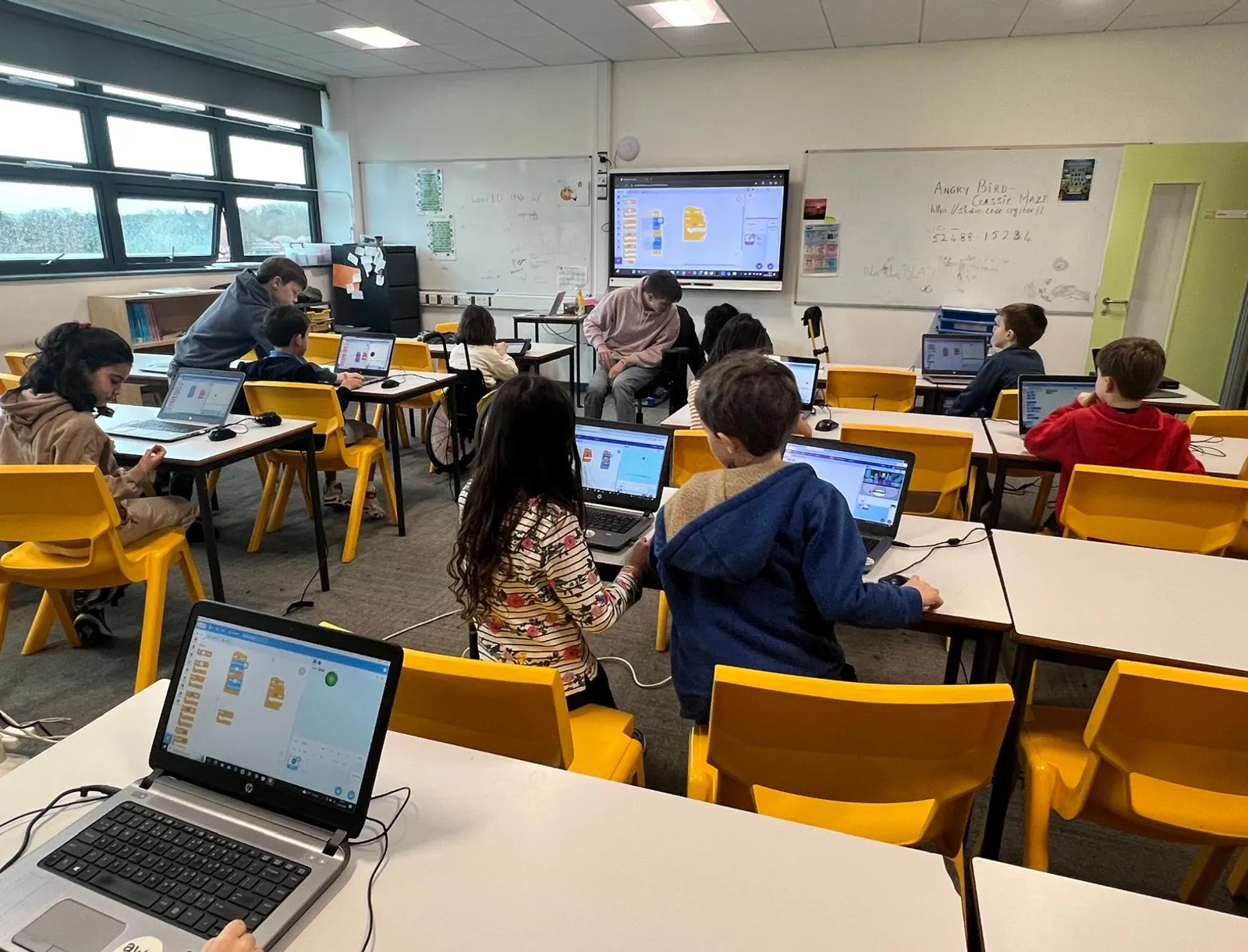 A classroom of children at laptops writing block code, with an instructor helping at the front and a project open on the interactive whiteboard